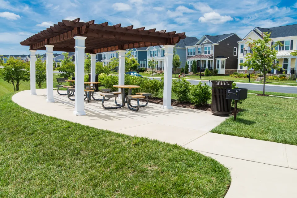 A pergola with a grill is located in a grassy park, across the street from single-family housing in a suburban style.